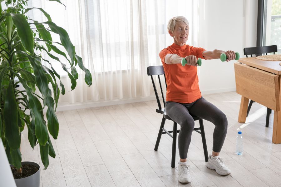 An older woman sits on a chair indoors, performing a seated strength exercise by lifting small green dumbbells in a bright, calm living space.