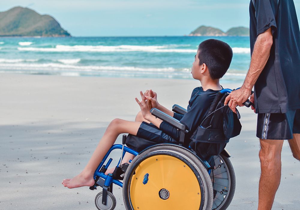 disabled boy in wheelchair pushed along beach with ocean in the background